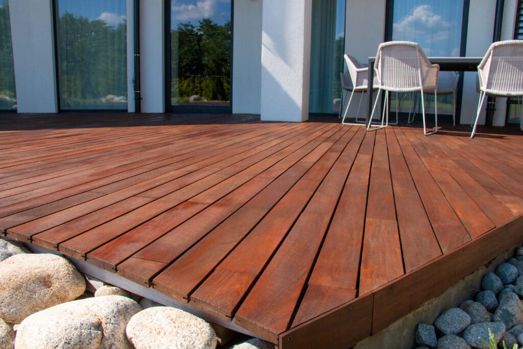 Grande terrasse en bois rouge avec salon de jardin extérieur devant une maison moderne aux baies vitrées. Bordure de galets.