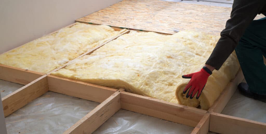 Un ouvrier en gant rouge pose de la laine minérale pour isoler un plancher. L'isolant est déroulé entre une ossature bois et sous des panneaux OSB.