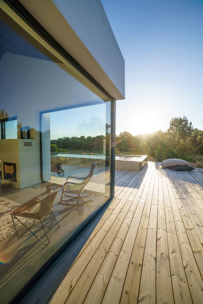 Maison moderne avec grande baie vitrée ouvrant sur une vaste terrasse en bois. Lumière dorée du soleil couchant sur le paysage boisé et l'eau.